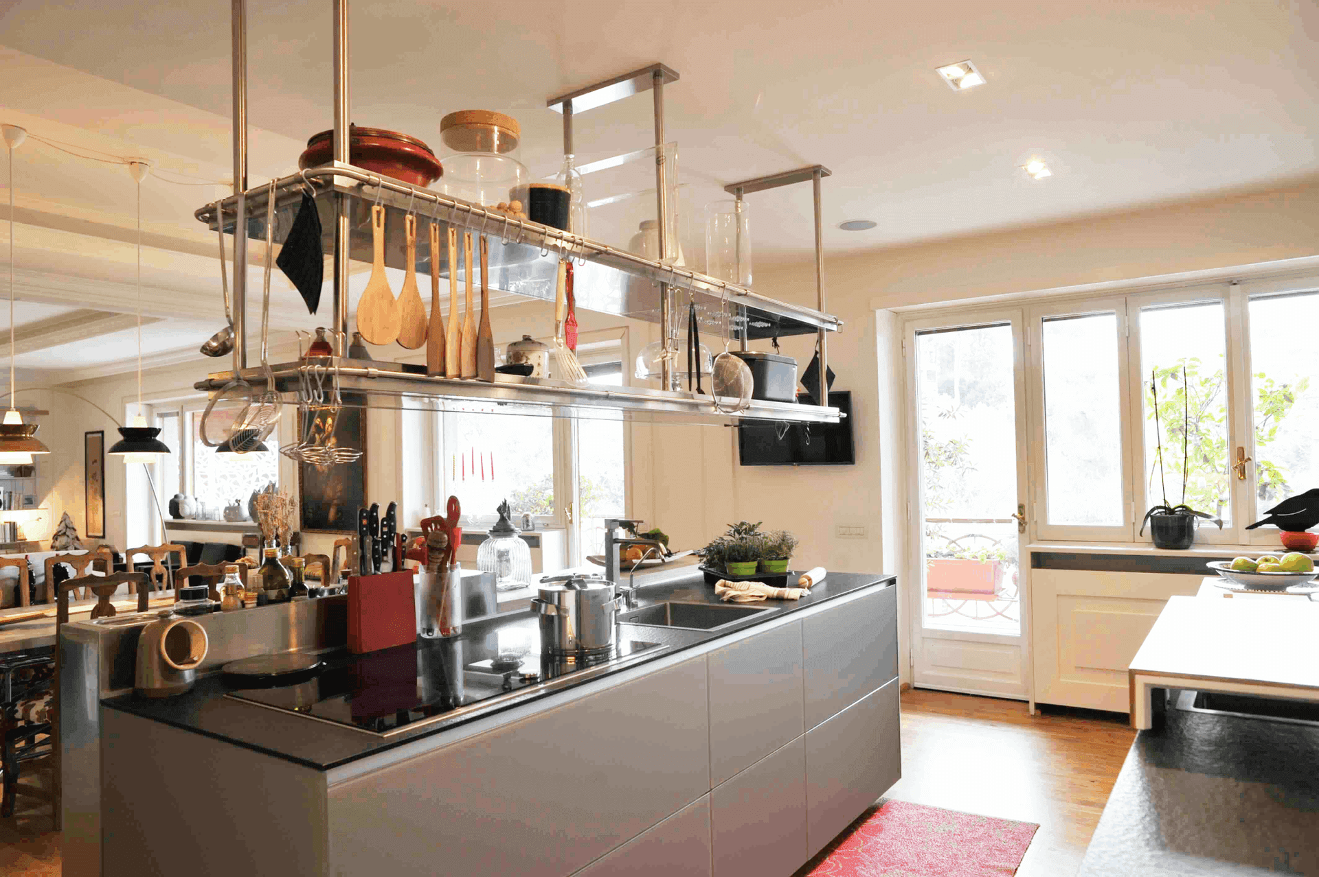 Modern kitchen with island, hanging utensils, large windows, and potted plants on the counter.